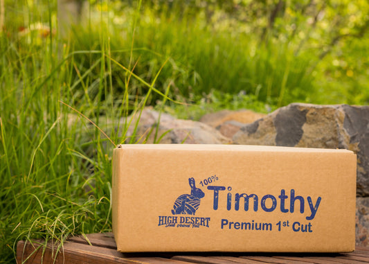 Cardboard box with 'Timothy High Desert' logo on a wooden surface with grass and rocks in the background