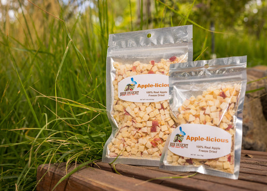 Two packages of Apple-licious dried apple pieces on a wooden surface with grass in the background